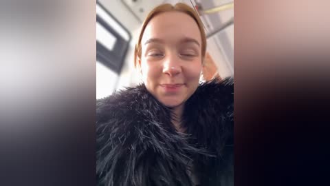 Media: A video of a smiling woman with light skin and blonde hair, wearing a black fur coat, taken from a low angle inside a bus.