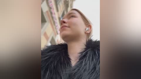 Media: A video of a fair-skinned, blonde woman with a slim build wearing a black fur coat and silver earrings, standing in front of a blurred, beige-colored building.