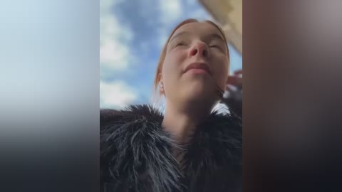 Media: Video of a fair-skinned, blonde woman with a serene expression, wearing a black fur coat, gazing upward at a bright blue sky with fluffy clouds.