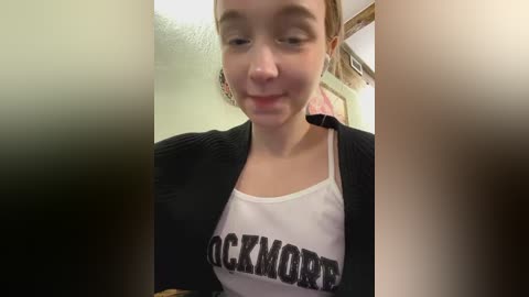 Media: A video of a young woman with light skin and brown hair, wearing a white tank top with \"BLOCKMORE\" printed in bold black letters. She has a slight smile and is indoors, partially obscured by out-of-focus objects in the foreground.