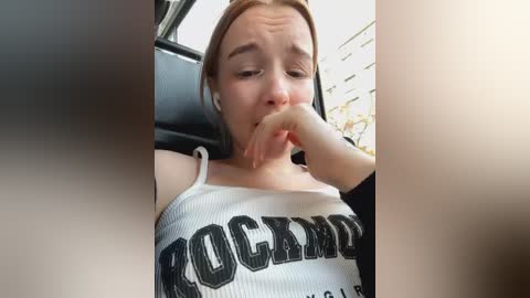 Media: Video of a young woman in a car, wearing a white \"ROCKANDROLL\" tank top, biting her finger, with blurred car interior and street outside.