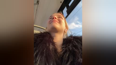 Media: Video of a blonde woman with fair skin, wearing a black fur coat, standing in a sunlit room with a large window showing a clear blue sky.