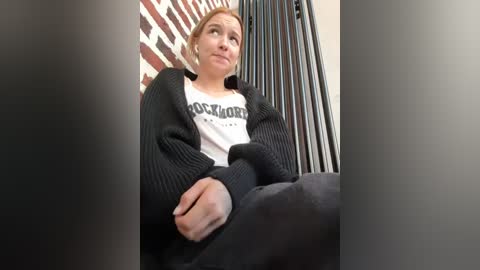 Media: Video of a fair-skinned, blonde woman with a neutral expression, wearing a black cardigan over a white \"Hooters\" shirt and black pants, seated in front of a brick wall and vertical blinds.