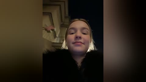 Media: Video of a young, light-skinned girl with shoulder-length blonde hair, wearing a black coat, standing in a dimly lit hallway with geometric wallpaper and a staircase in the background.