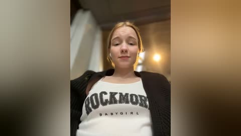 Media: Video of a young Caucasian woman with light skin and blonde hair, wearing a white babydoll top with \"ROCKAMOR BABY\" text, black knit cardigan, and neutral background.