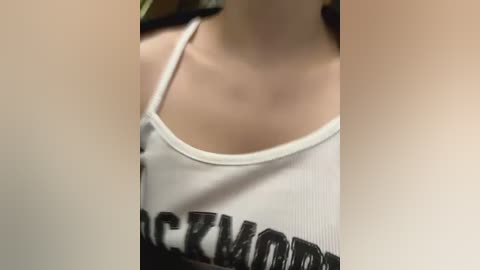 Media: A close-up video of a woman in a white tank top with black \"ROCKWOOD\" text, her bare shoulders and cleavage visible, necklaces hanging. The background is blurred, focusing on the top.