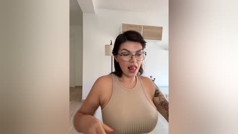 Media: Video of a woman with glasses, medium build, and medium-sized breasts, wearing a sheer beige top, posing indoors in a modern, minimalist room with white walls and light wooden floors.