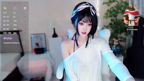 Media: Video of an Asian woman with black hair and a white headband, wearing a white off-shoulder top, holding a controller in a modern living room with a TV, plant, and furniture.