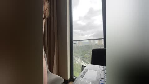 Media: Video of a person with long blonde hair, seated at a desk with a laptop, looking out a large window at a cloudy sky and distant cityscape.