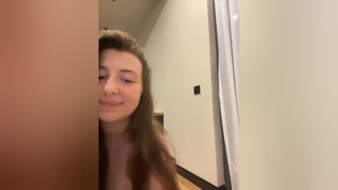 Media: Video of a young woman with long brown hair, light skin, and a slim build, smiling softly while partially obscured by a mirror in a modern, minimalist room with beige walls and a wooden floor.