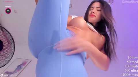 Media: Video of a Latina woman with long black hair, medium skin tone, and large breasts, performing a high kick in tight blue leggings, in a dimly lit room.