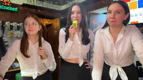 Media: Video of three young women, two with long hair, wearing white blouses tied at the waist and black pants, standing indoors near a bar, holding a green phone, smiling, casual attire.