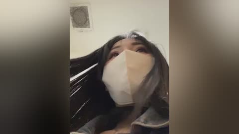 Media: Video of a person wearing a white surgical mask, with long dark hair, inside a dimly lit room with a metal vent on the wall.