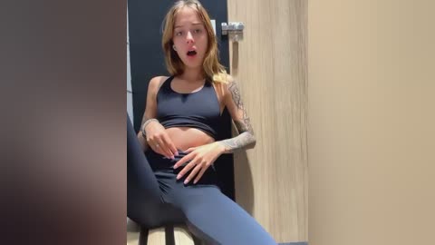 Media: Video of a slim, tattooed blonde woman in a black sports bra and leggings, sitting in a changing room, hand on her belly, mouth open in surprise.