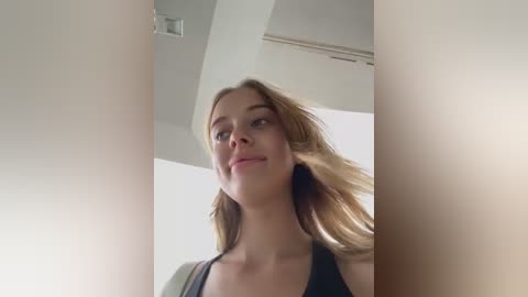 Media: A video of a young woman with fair skin and long blonde hair, wearing a black top, smiling confidently, taken from a low angle, with a bright, minimalist background.