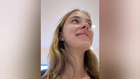 Media: Video of a young Caucasian woman with light brown hair, looking upward with a thoughtful expression, blurred background, indoors.