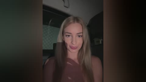 Media: Video of a blonde woman with light skin and long hair, wearing a sheer, burgundy top, sitting in a dimly lit car, with blurred background.