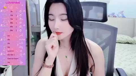 Media: Video of an East Asian woman with long black hair, fair skin, and a slim physique, wearing a sleeveless top, sitting in a gray office chair. She has a contemplative expression, touching her chin. Background includes a pink wall calendar and a white fluffy rug.