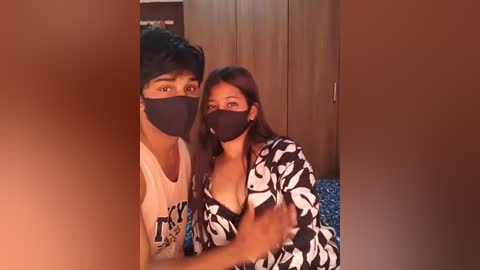 Media: Video of a young couple wearing black face masks, with the man in a white T-shirt and the woman in a black-and-white floral dress, standing indoors near a wooden wardrobe.