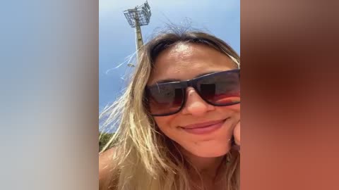 Media: Video of a smiling, tanned woman with long, blonde hair, wearing dark sunglasses and a red swimsuit, standing in front of a light blue sky with a basketball hoop in the background.