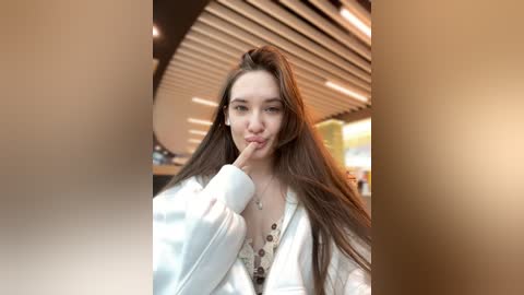 Media: Video of a young woman with long brown hair, wearing a white jacket, and holding a straw to her mouth, set in a modern, brightly lit restaurant with wooden ceiling panels.