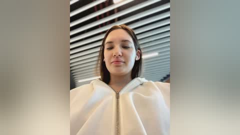 Media: Video of a young woman with straight brown hair, wearing a white zip-up jacket, standing in front of horizontal blinds. She has a neutral expression, and the background is softly blurred.