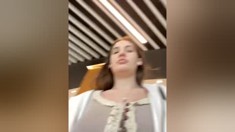 Media: A blurry video of a young woman with light skin, brown hair, wearing a white cardigan over a light-colored blouse, standing in a modern indoor setting with a wooden ceiling and ambient lighting.