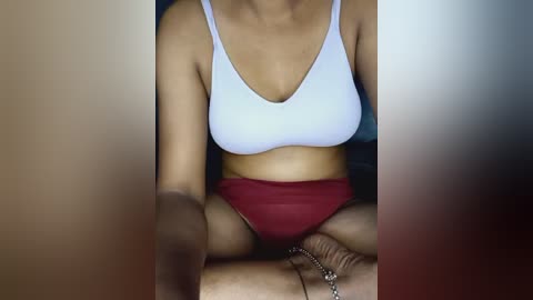 Media: A video of a woman with medium brown skin, wearing a white sports bra and red shorts, sitting cross-legged on a wooden floor, with a blurred background.