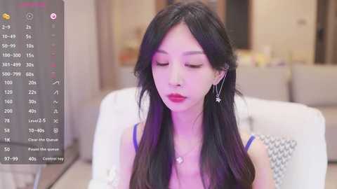 Media: Video of an East Asian woman with long black hair, wearing a blue top, sitting on a white couch in a modern living room.