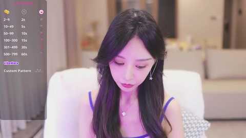 Media: A video of an Asian woman with long black hair, wearing a blue dress, sitting indoors, with a blurred background showing a beige sofa.