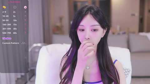 Media: Video of an East Asian woman with long black hair, wearing a purple bra, biting her finger, in a modern living room with white furniture.