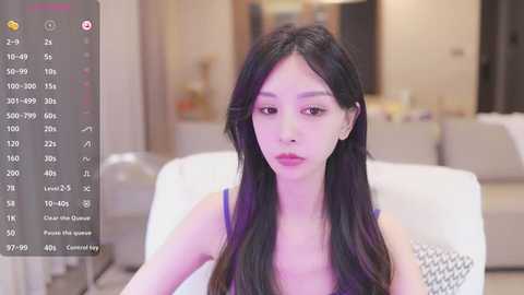 Media: Video of a young Asian woman with long black hair and fair skin, wearing a purple top, seated indoors on a white couch. Background includes blurred modern living room decor.