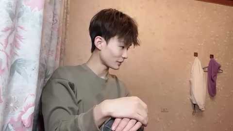 Media: Video of a young East Asian man with short dark hair in a green shirt, standing in a bathroom with floral-patterned curtains, beige wallpaper, and a towel rack holding purple and white towels.