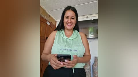 Media: Video of a smiling Latina woman with long black hair, wearing a green sports tank top and black pants, holding a smartphone. Background includes a wooden door, ceiling fan, and a TV.