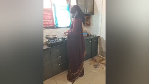 Media: Video of a woman in a maroon sari with a white border, cooking in a small, dimly lit kitchen with white tiles and dark wooden cabinets.