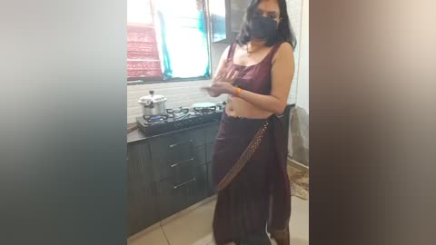 Media: Video of a woman in maroon saree and face mask, cooking on a stove in a small, cluttered kitchen with tiled walls.