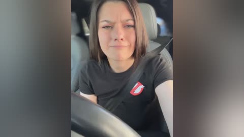 Media: Video of a young woman with shoulder-length brown hair, wearing a black t-shirt with a red and white logo, sitting in a car with a neutral expression.