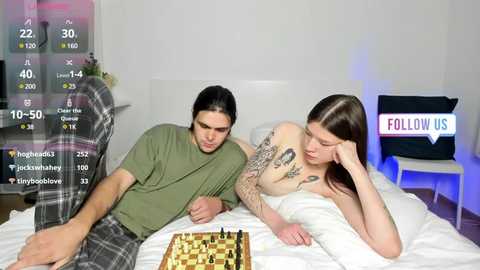 Media: A video of a man and woman in a bedroom, playing chess on a bed. The man has long hair, wears a green shirt, and plaid pajama pants. The woman is topless, with tattoos, lying on her side.