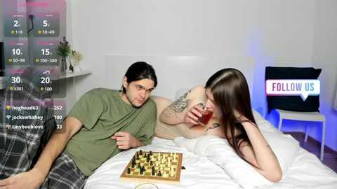 Media: Video of a young couple playing chess on a bed. Man with long hair in a green shirt, woman with long brown hair, tattooed arm.