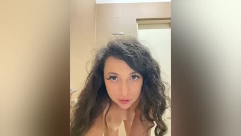 Media: Video of a nude, fair-skinned woman with long, curly black hair and large breasts, standing in a beige-tiled bathroom, looking directly at the camera.