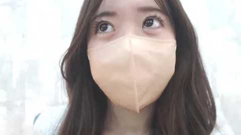 Media: Video of an Asian woman with long brown hair, wearing a beige surgical mask, looking upwards with wide, expressive eyes. Background is blurred, likely an indoor setting with light colors.