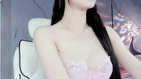 Media: Video of a young, fair-skinned woman with long black hair, wearing a delicate pink lace bra with thin straps, seated on a white, cushioned chair against a gray marble wall with a blurred, abstract artwork in the background.