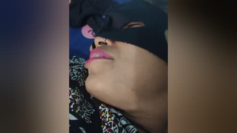 Media: Video of a person lying down with a black blindfold over their eyes, revealing pink lips and a nose ring. They wear a floral-patterned shirt, with a dark background.