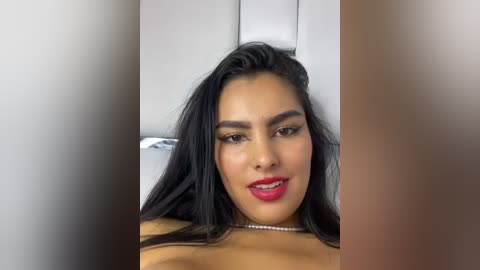 Media: Video of a Latina woman with long, black hair, wearing bold red lipstick, and a beaded necklace, lying on a white bed, with blurred faces in the background.