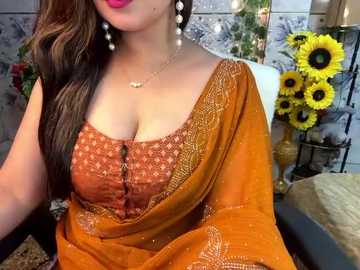 Media: Video of a woman with fair skin, wearing an orange saree with intricate embroidery, adorned with pearl earrings, seated in a cozy indoor setting with sunflowers and a white cushion in the background.