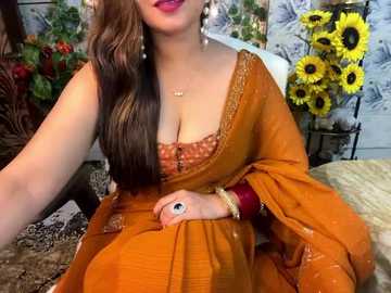 Media: Video of a South Asian woman in an orange sari with a low neckline, adorned with gold jewelry and a flower on her wrist. Background features sunflowers and greenery.