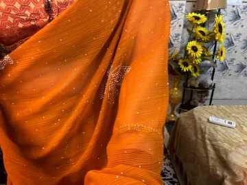 Media: Video of an orange saree with gold sequins, draped over a person, in a room with floral wallpaper, a sunflower arrangement, and a beige bedspread.