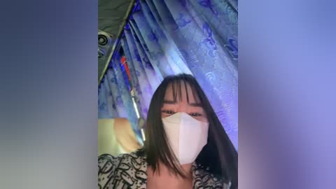 Media: A video of an Asian woman with shoulder-length dark hair, wearing a white surgical mask, standing in a dimly lit room with blue and purple curtains.