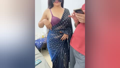 Media: A video of a plus-sized woman in a dark blue sari with white stripes, revealing a purple bra underneath, smiling and adjusting her sari. A man in a red shirt is partially visible on the right.