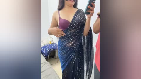 Media: Video of a South Asian woman in a purple bra, holding a phone, draped in a blue sari with white lines. Background shows a messy bedroom with a bed, wall, and red shirt.